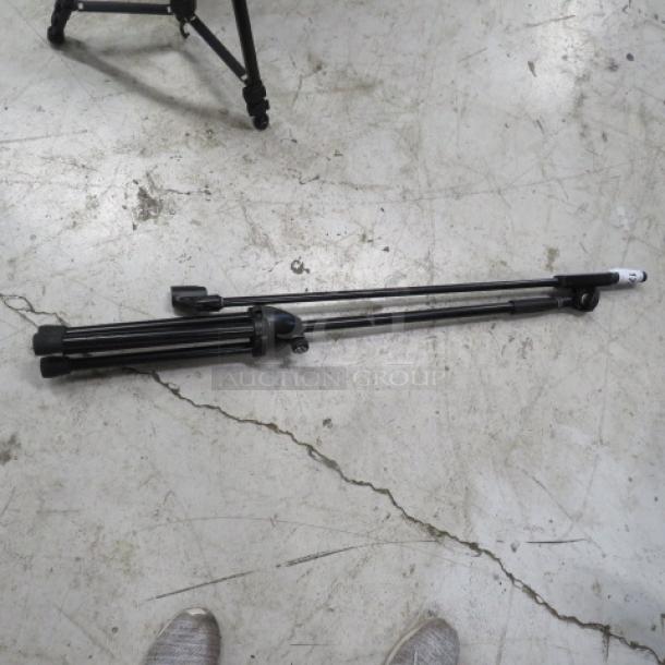 Collapsible black mic stand on concrete floor, folded. Good condition, with adjustable legs and tripod base.