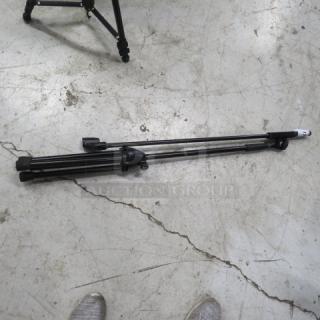 Collapsible black mic stand on concrete floor, folded. Good condition, with adjustable legs and tripod base.