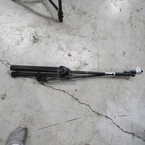 Collapsible mic stand, black metal, folded, good condition. Features adjustable legs and a lock mechanism. No visible brand marks.
