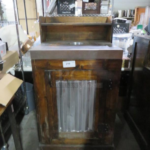 Wooden and metal trash receptacle with stainless steel top, includes trash can. Features a small shelf and numbered tag "210".