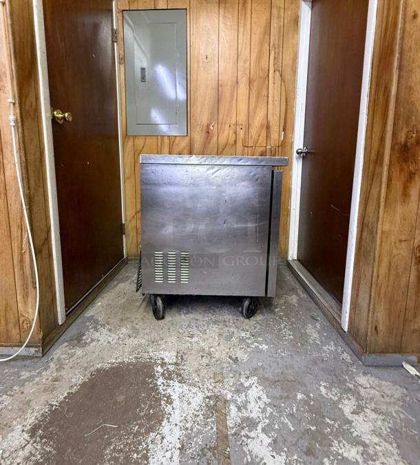 Asbury Arctic Air MCF-27U 27" undercounter freezer, stainless steel, used condition, on casters, in wood-paneled room.