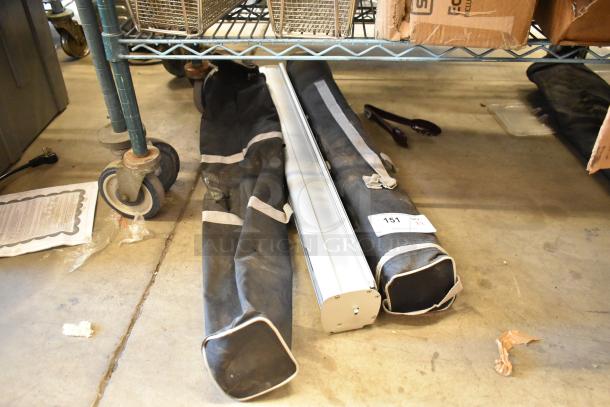 Two black soft bags with gray straps, containing a retractable easel. Label states "151, Qty x2". Placed on a concrete floor.