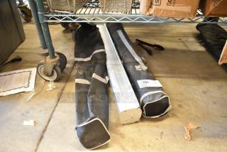 Two black soft bags with gray straps, containing a retractable easel. Label states "151, Qty x2". Placed on a concrete floor.