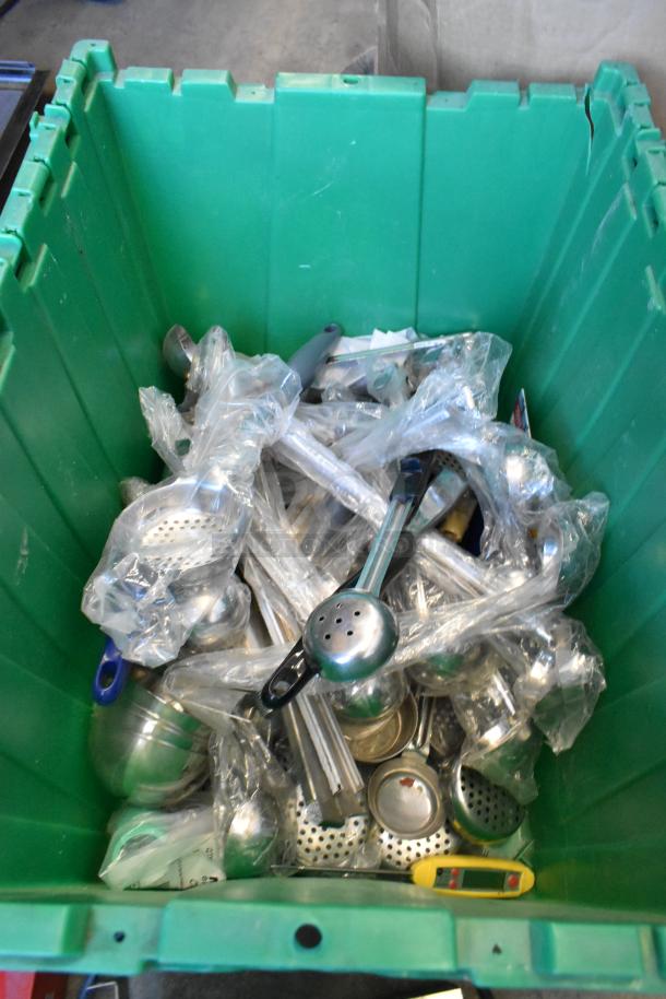 Green poly bin filled with various metal items wrapped in plastic, including strainers and a yellow device. Auction sale.