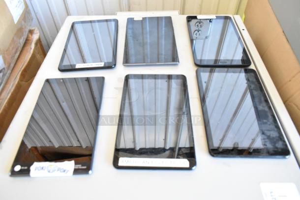 Six various tablets including Samsung, in good condition, displayed on a table. Some have labels with serial numbers.