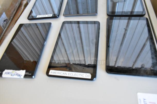 Six various tablets on a table, including Samsung models, with visible reflection and labels. Tablets are in used condition.