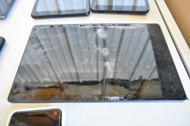 Six assorted tablets on a table, one with a cracked screen. Various models and conditions. No visible tags or labels.