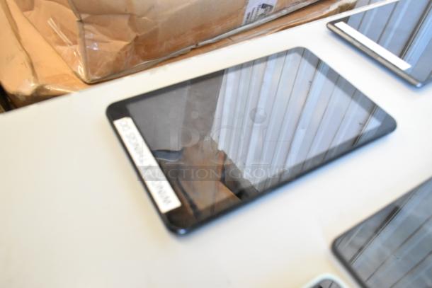 Six various tablets on a table, each with a reflective screen. One tablet shows a label on its edge. Condition appears used.