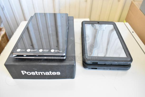 Five Samsung and Lenovo tablets, mixed models, on a table. Some show wear. Postmates box included. Features visible labels.