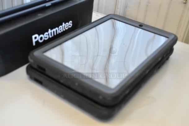 Five assorted Samsung and Lenovo tablets stacked, in black cases. "Postmates" box in background. Condition appears used.
