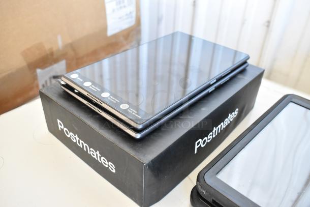 Five Samsung and Lenovo tablets stacked on a Postmates box. Features and condition vary. Minimal visible wear.