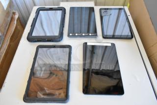 Five various tablets including Samsung, displayed on a white surface. Some devices have black protective cases. Appears in good condition.