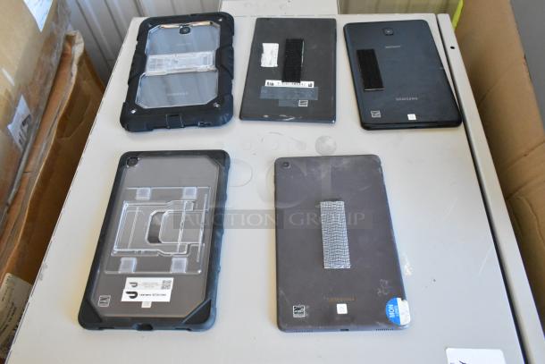 Five tablets including Samsung models, mixed conditions, some with protective cases, visible labels and attachments.