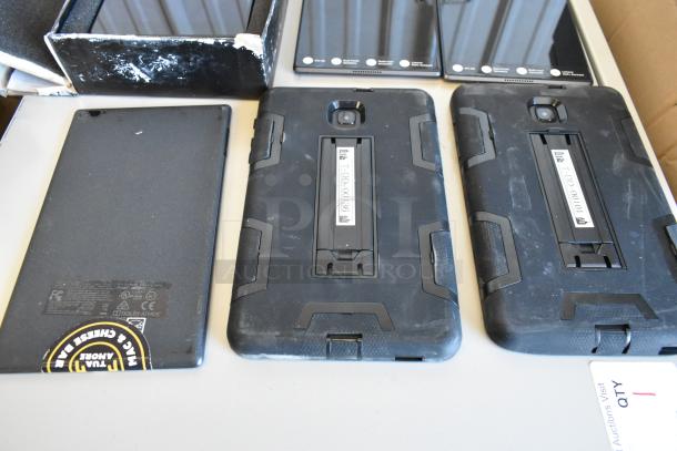 Six assorted tablets, including Samsung, in used condition. Each has a protective black case. Some wear on cases visible.