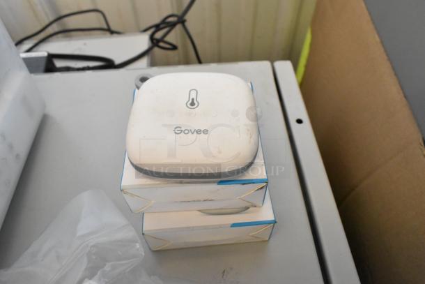 Lot of various items, including a Govee Wi-Fi thermo-hygrometer, visible on a table. Hygrometer shows minor wear.