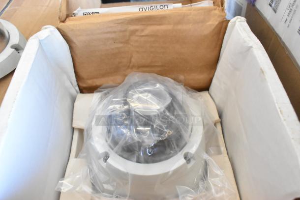 Brand new Avigilon camera in original packaging, covered in protective plastic. Box includes manuals and installation materials.