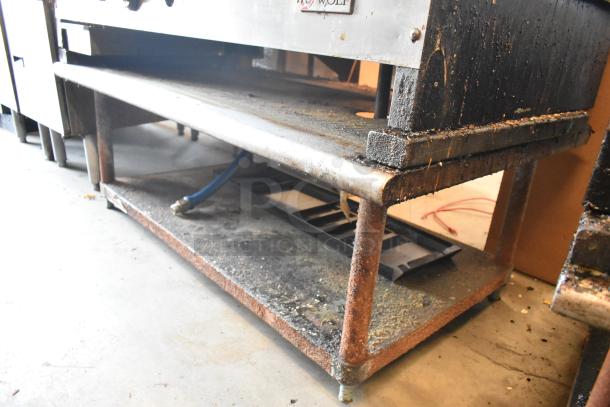 Wolf stainless steel commercial gas griddle on metal stand with under shelf, showing wear and attached blue gas hose.