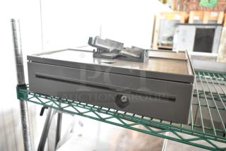 Metal cash drawer with lock, keyhole visible, and a flat top. Located on a green metal shelf. In good condition.