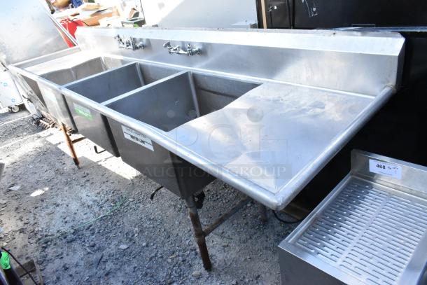 Stainless steel 3 bay sink with dual drain boards, visible faucets, and sturdy construction. Slight wear, ideal for commercial use.