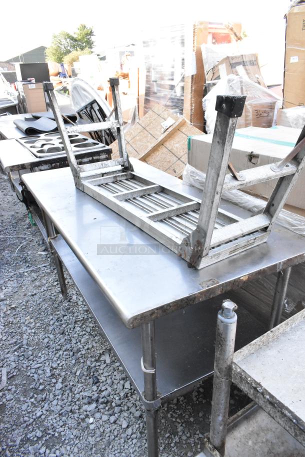 Stainless steel table with under shelf, shown outdoors. Features tubular legs, brushed finish, slight wear visible.