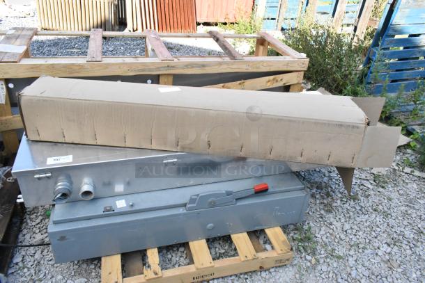 Metal power box with levers and two additional items, including a cardboard box, on a wooden pallet.