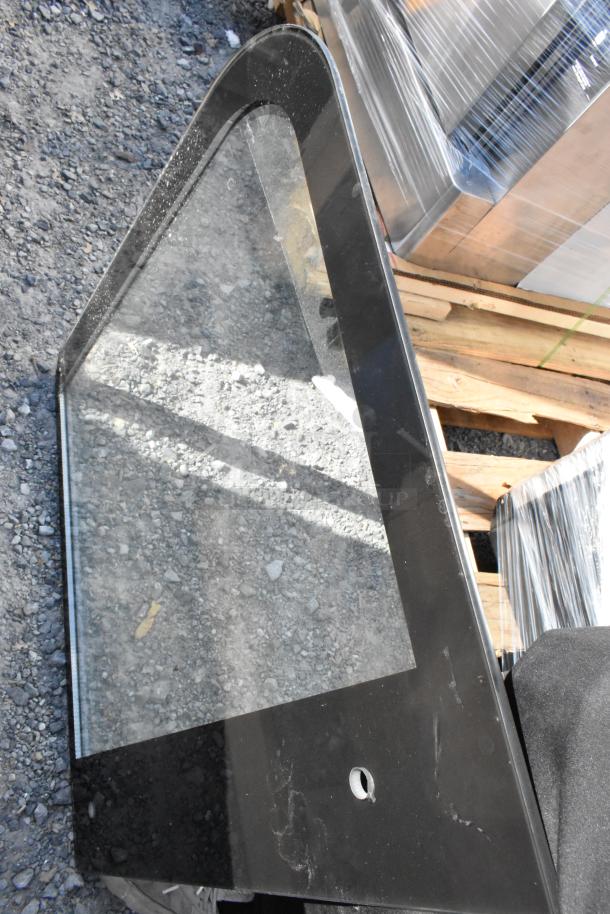 Acrylic panel with black frame, showing light scratches, resting on gravel next to pallet with wrapped items.