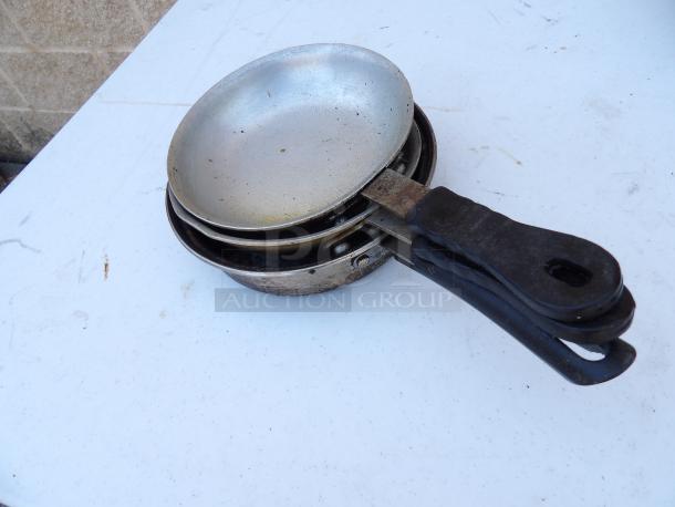 Set of three 7" commercial frying pans with rubber grips, showing visible wear, stacked on a white surface.