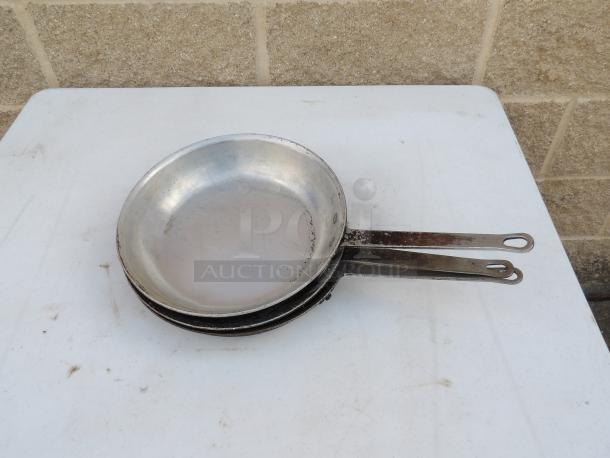 Set of three 10" aluminum fry pans, pre-owned, showing signs of wear. Sturdy metal handles with hanging holes.