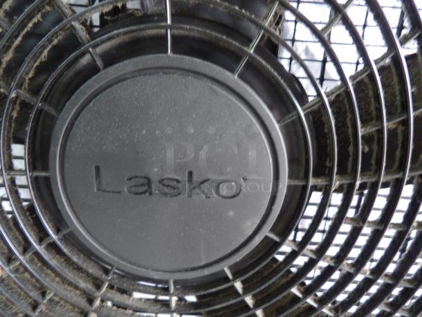 Lasko box fan, visible dust accumulation, black plastic guard, model powers on, brand name engraved on center.