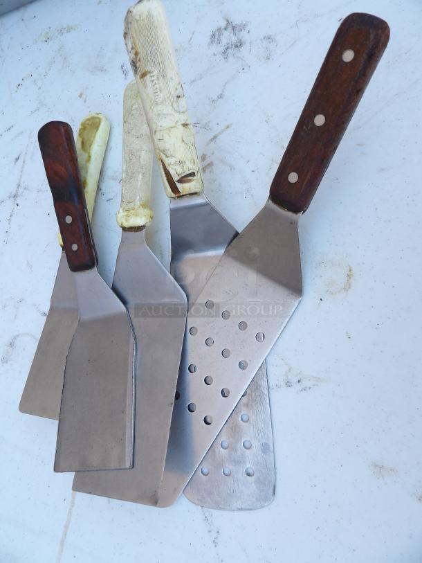 Set of five metal spatulas with wooden and plastic handles, showing signs of wear, one with perforated blade.