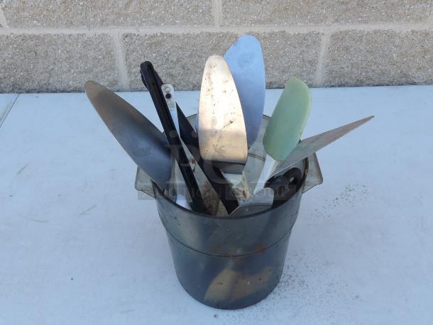 Miscellaneous pie spatulas in a metal container, various sizes and conditions, suitable for kitchen use.