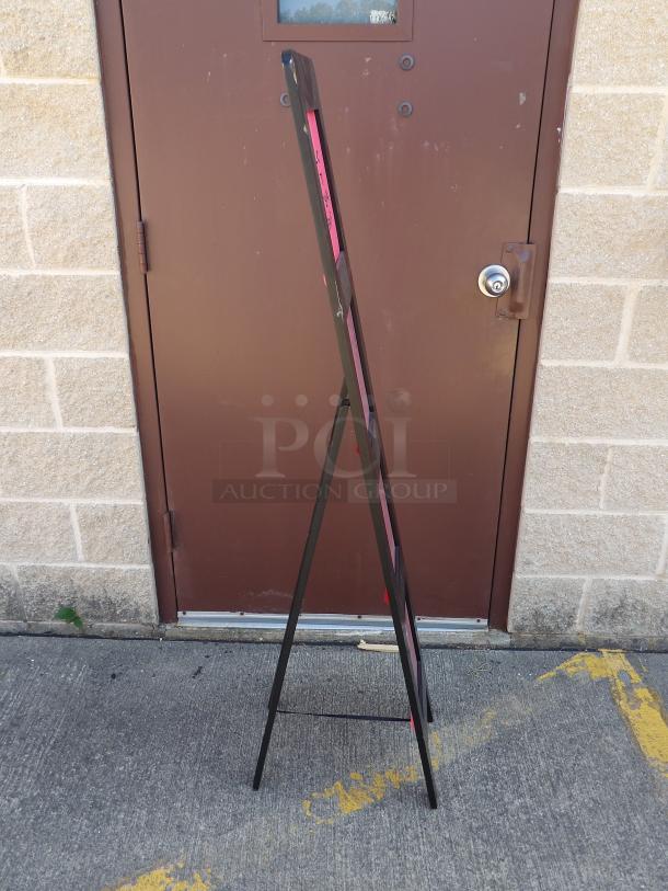 Wooden display sign easel, black finish, sturdy build, slight wear visible, standing against a brick wall.