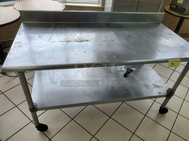 Stainless steel table with a backsplash and lower shelf, showing signs of wear. Equipped with caster wheels for mobility.
