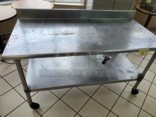 Stainless steel table with a backsplash and lower shelf, showing signs of wear. Equipped with caster wheels for mobility.