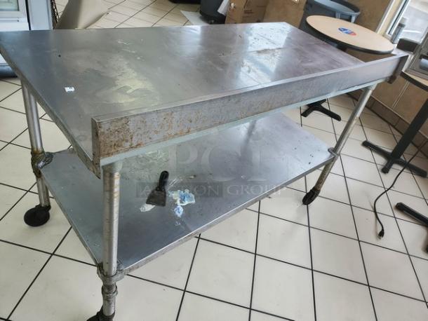 Stainless steel table with two shelves, showing signs of wear and rust. Features caster wheels for mobility.
