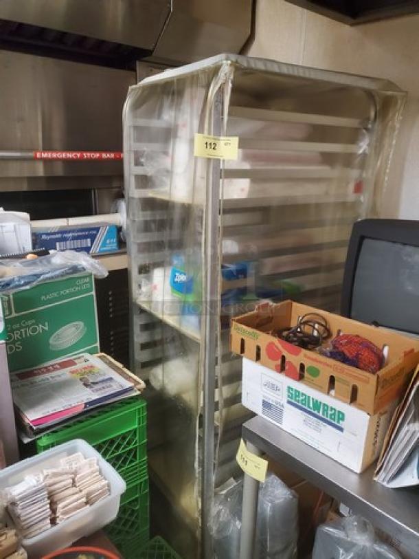 Stainless steel sheet pan rack with multiple shelves, used condition, clear cover, tag marked "112." Contents not included.