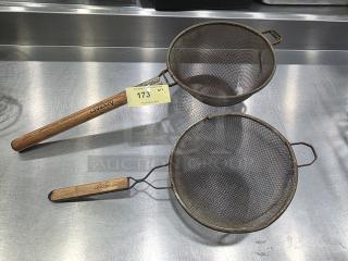 Two metal strainers with wooden handles on stainless steel surface, marked "2XBID" and "173." Slight rust visible.