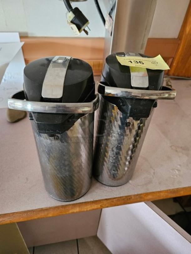 Lot of two Newco Thermal Airpots in stainless steel. Features carry handles. Minor wear visible. Ideal for hot beverages.