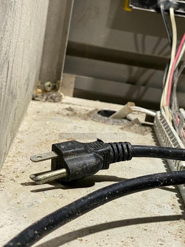 A black electrical plug connected to a dusty power cord lies on a rough, industrial surface near other wires.