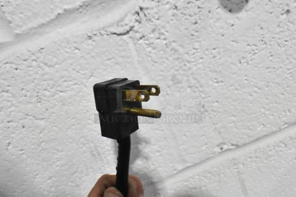 Close-up of a three-prong plug from a Beverage Air SMF58HC-1-S milk cooler, 115 volts, 1 phase, against a textured wall.