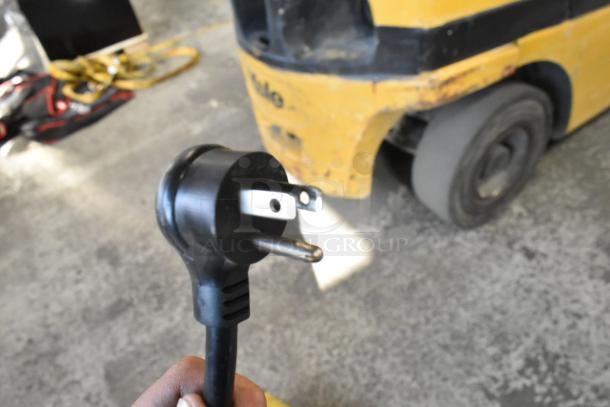 Close-up of a three-prong electrical plug, blurred background with a yellow vehicle, unrelated to the described auction item.