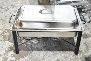 Brand new chafing dish with minor scratches, stainless steel, rectangular with lid and handles, stands on black metal legs.