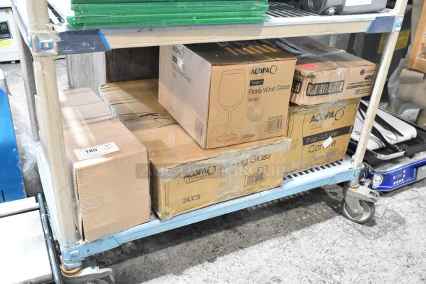 Boxes of Acopa and Anchor Hocking glassware on a cart, including wine glasses, mixing glasses, and carafes. New condition.