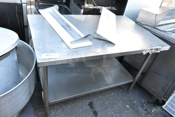 Stainless steel table with under shelf, industrial grade, includes two mounted tools. Slight wear visible, ideal for storage.