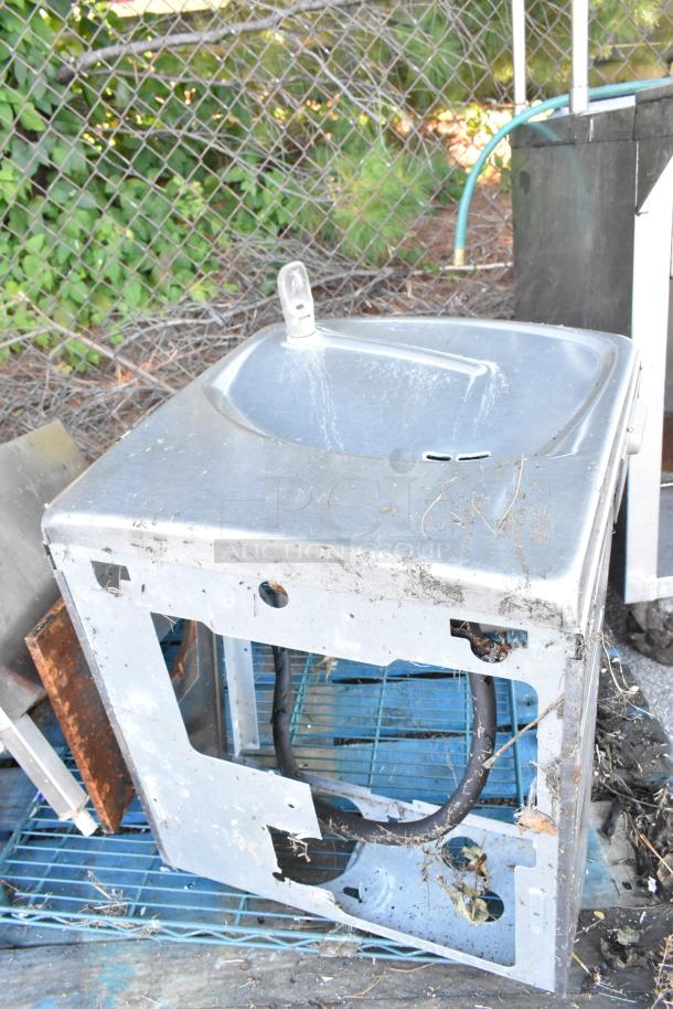 Metal wall mount water fountain, visibly worn with dirt and rust, missing panel, outdoor setting. Auction item.