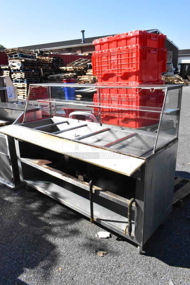 Stainless steel commercial steam table with sneeze guard and under shelf, moderate wear, outdoor setting.