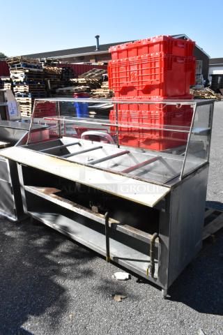 Stainless steel commercial steam table with sneeze guard and under shelf, moderate wear, outdoor setting.