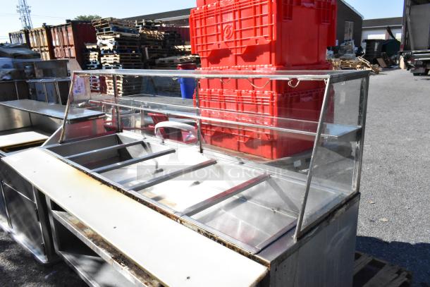 Stainless steel commercial steam table with sneeze guard and under shelf, visible wear, ideal for food service use.