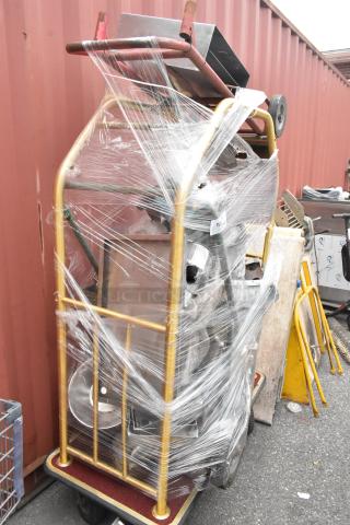 Metal luggage cart on commercial casters wrapped in plastic, containing metal baking pans and a poly trash can.