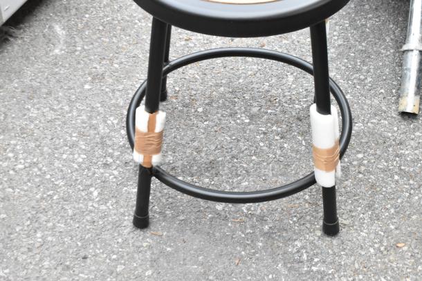 Brand new black metal stool with round footrest, showing protective padding on legs. Scratch and dent condition.